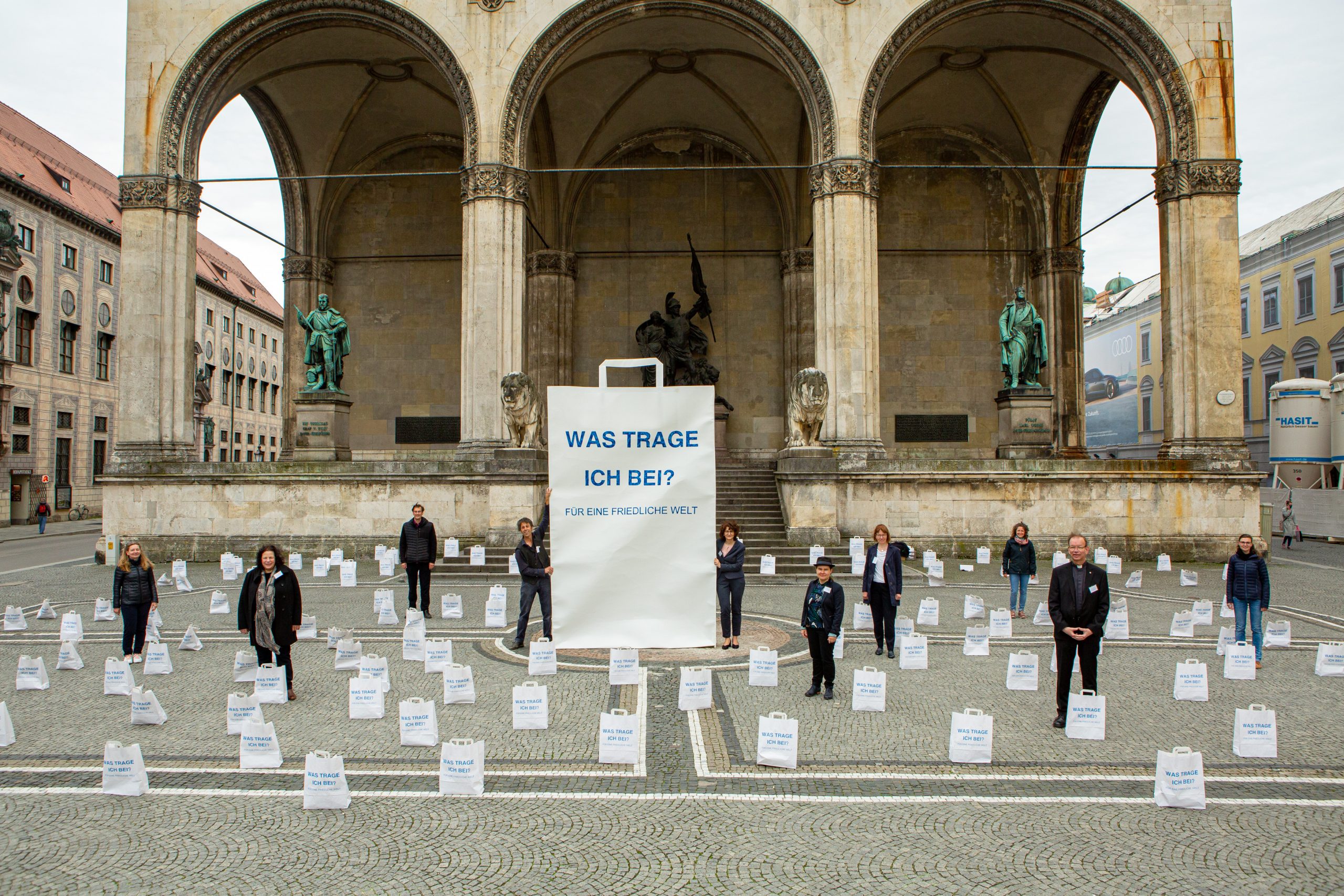 Eine Kunstinstallation aus Papier vor der Feldherrenhalle in München. Menschen stehen in einem Meer aus Papiertüten. In der MItte steht eine dreieinhalb Meter hohe Papiertüte. Alle sind bedruckt mit der Frage Was trage ich bei? Für eine friedliche Welt