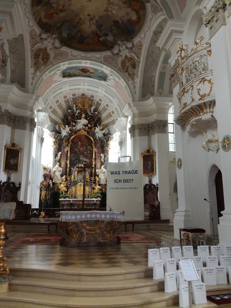 Eine 3,5m hohe Papiertüte steht gemeinsam mit kleinen aufgereihten Papiertüten in einer Kirche neben einem barocken Altar.